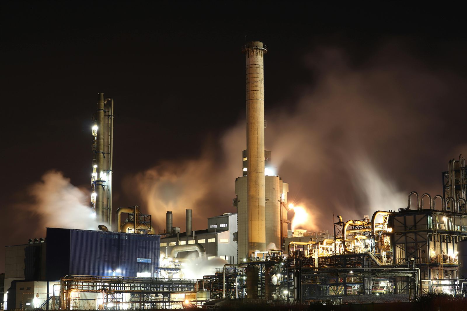 Planta petroquímica industrial operando de noche con chimeneas, vapor y luces, monitoreada con IA para gestión de riesgos HSE